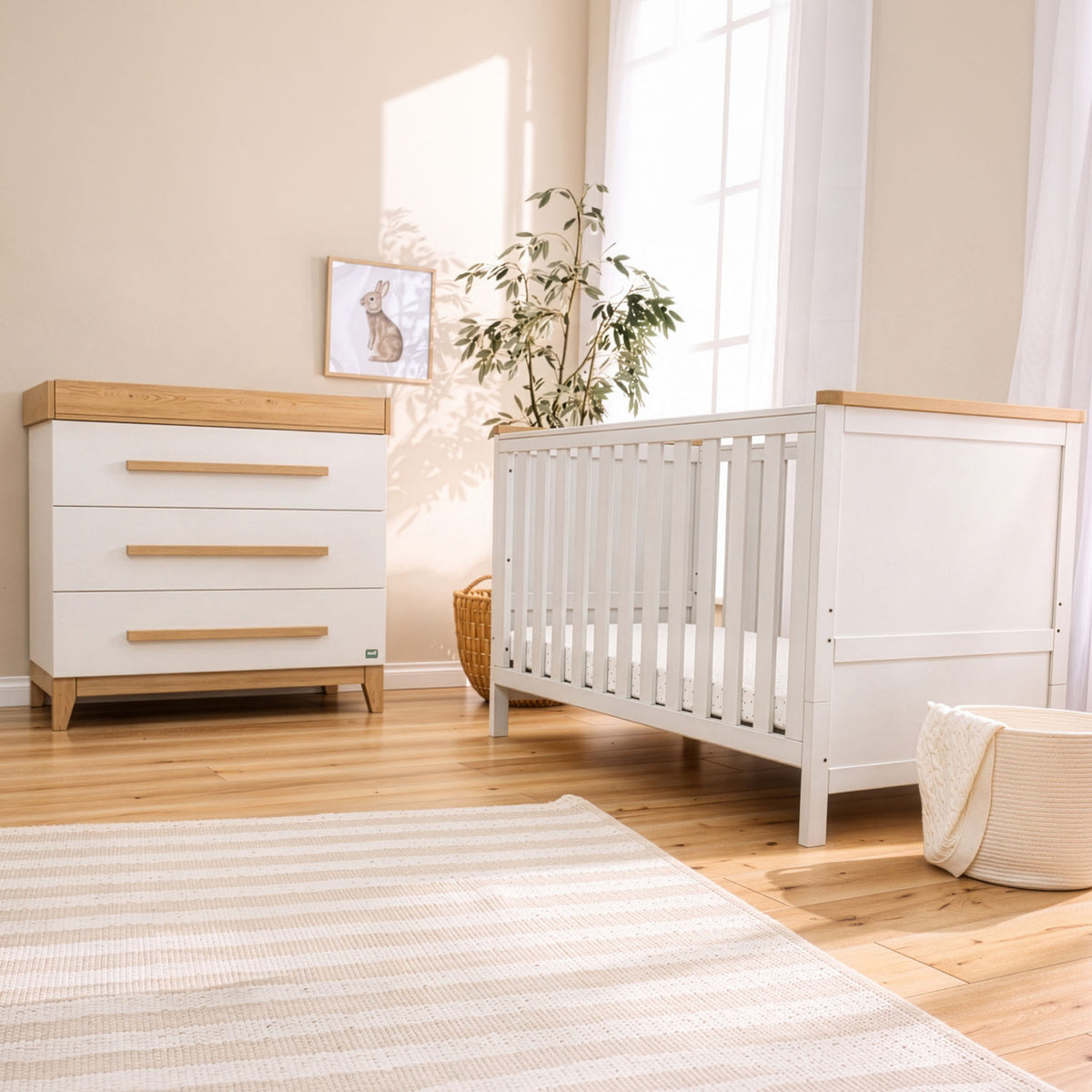 A white and natural golden oak baby nursery furniture set, including a chest of drawers and a cot bed, styled in a bright, sunlit nursery with a striped rug, wooden floor, and a potted plant.