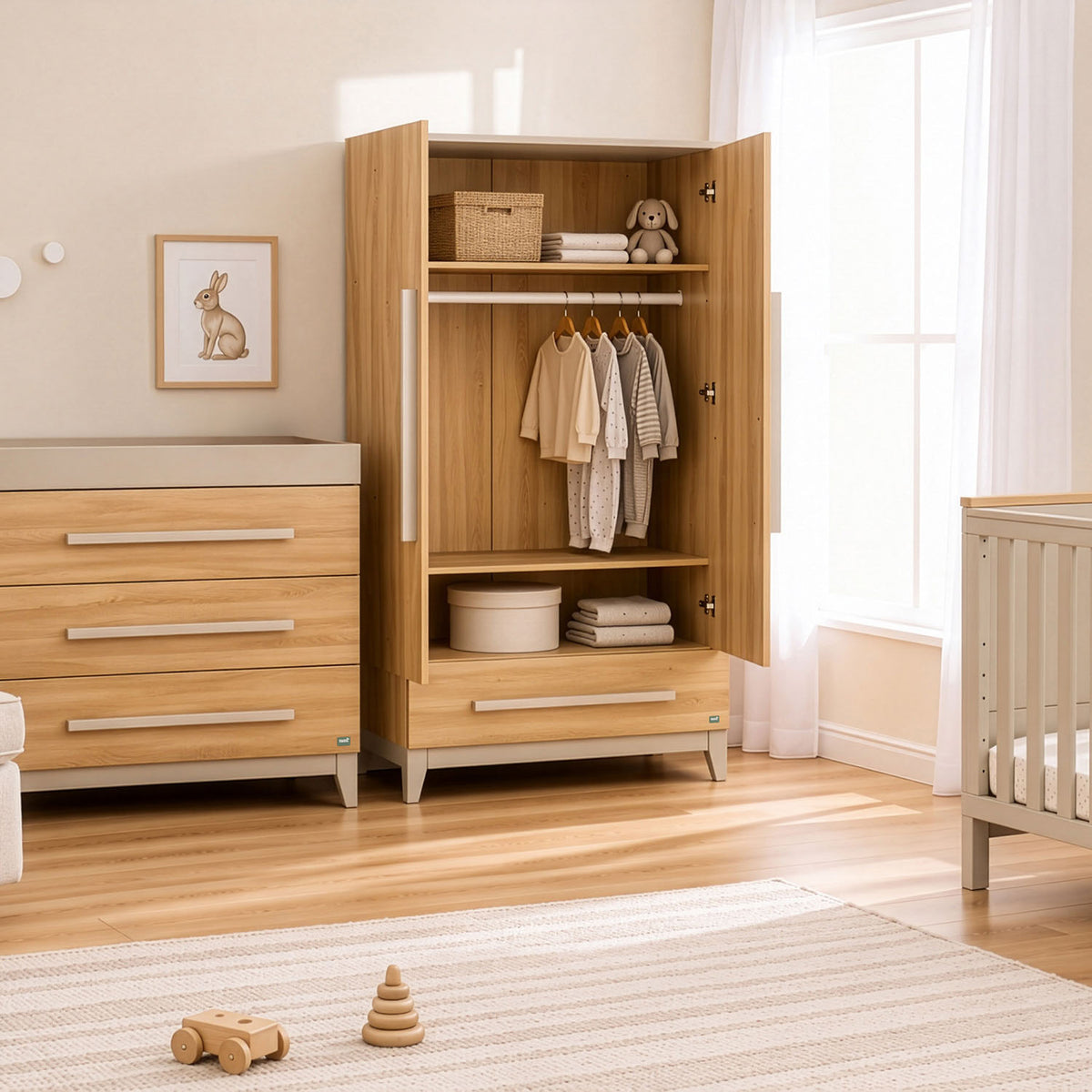 A light natural golden oak wood nursery wardrobe with open doors, styled in a bright nursery next to a matching wooden chest of drawers and cot bed.