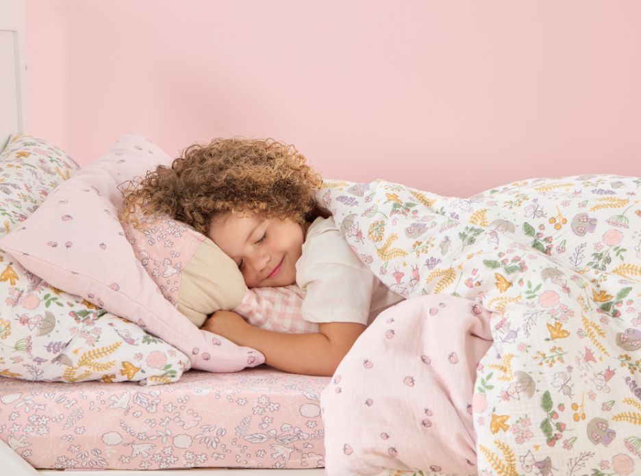 Curly-haired child cuddling under floral duvet with pink fitted sheet and matching mushroom cushion in cosy nursery.