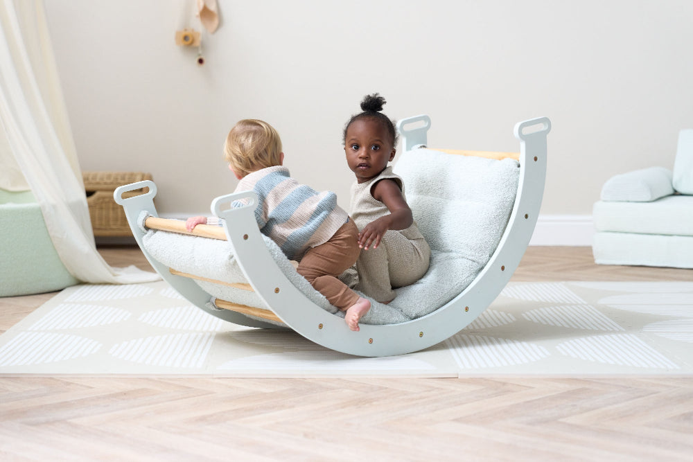 Two children sitting in a play arch in a room with a light-colored floor and white walls.