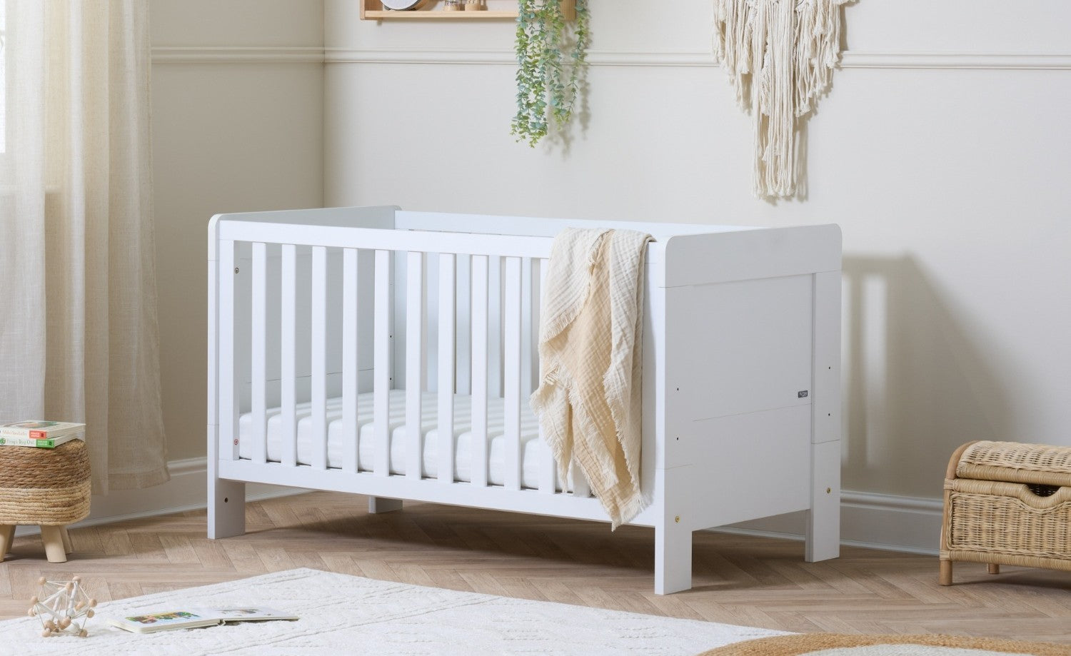 Alba cot bed in white with slatted sides, styled in a softly lit nursery with neutral accents, wooden flooring, and a light woven throw