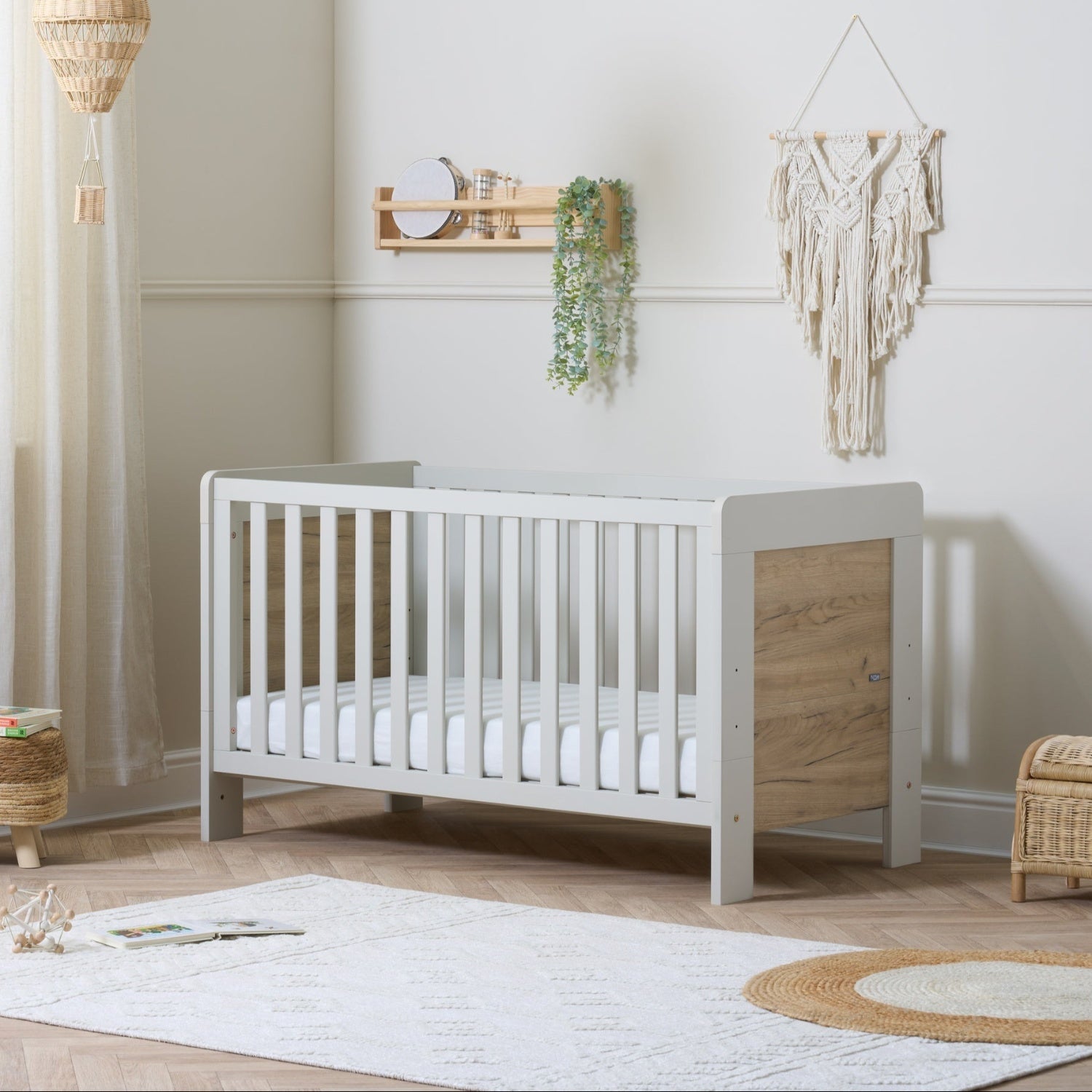 The image shows a modern Scandinavian-inspired cot bed with a white frame and distressed oak finish. It is set in a room with a light interior, and there are decorative items and a baby mobile above it.