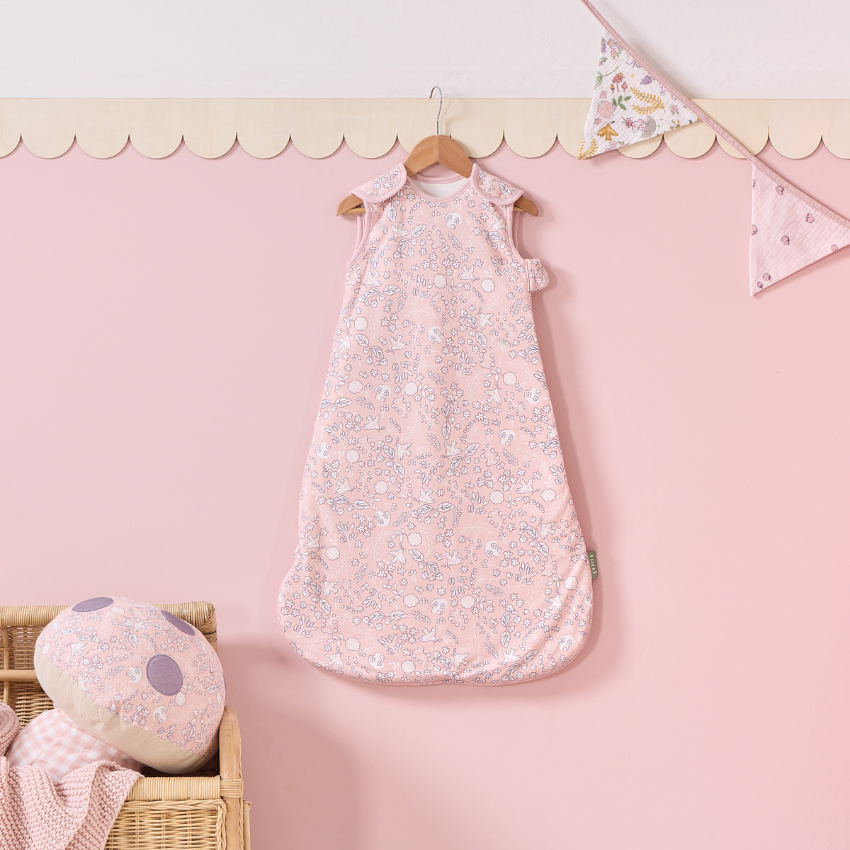 Tiny Tails baby sleep bag hanging on pink wall with bunting, styled with matching mushroom cushion and woven basket detail