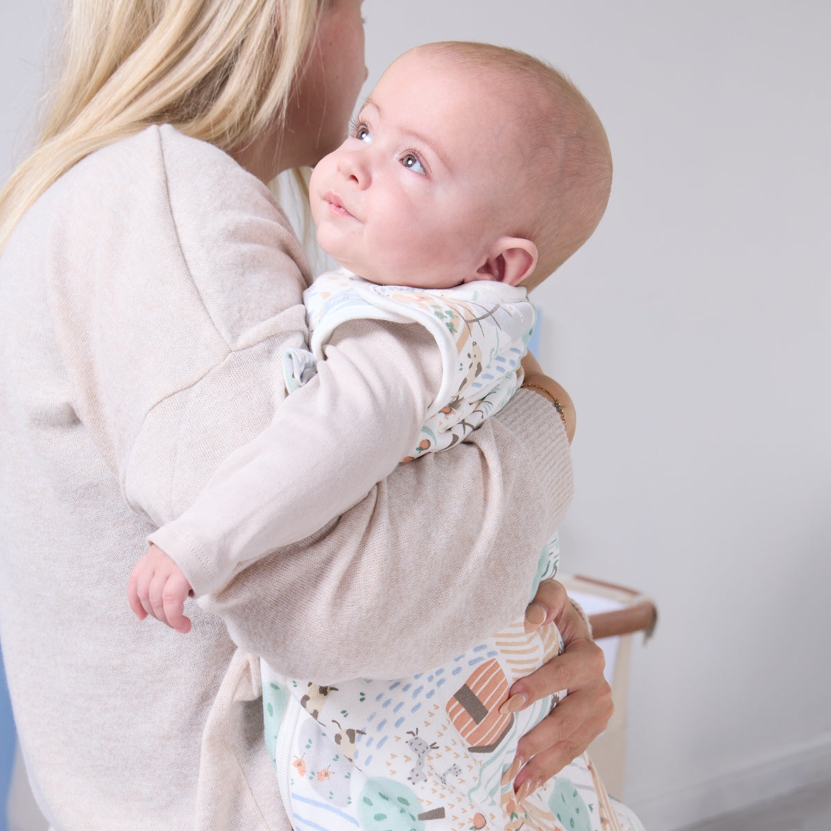 Mother holding baby dressed in the Good Life baby sleep bag, showing snug fit and soft pastel countryside pattern