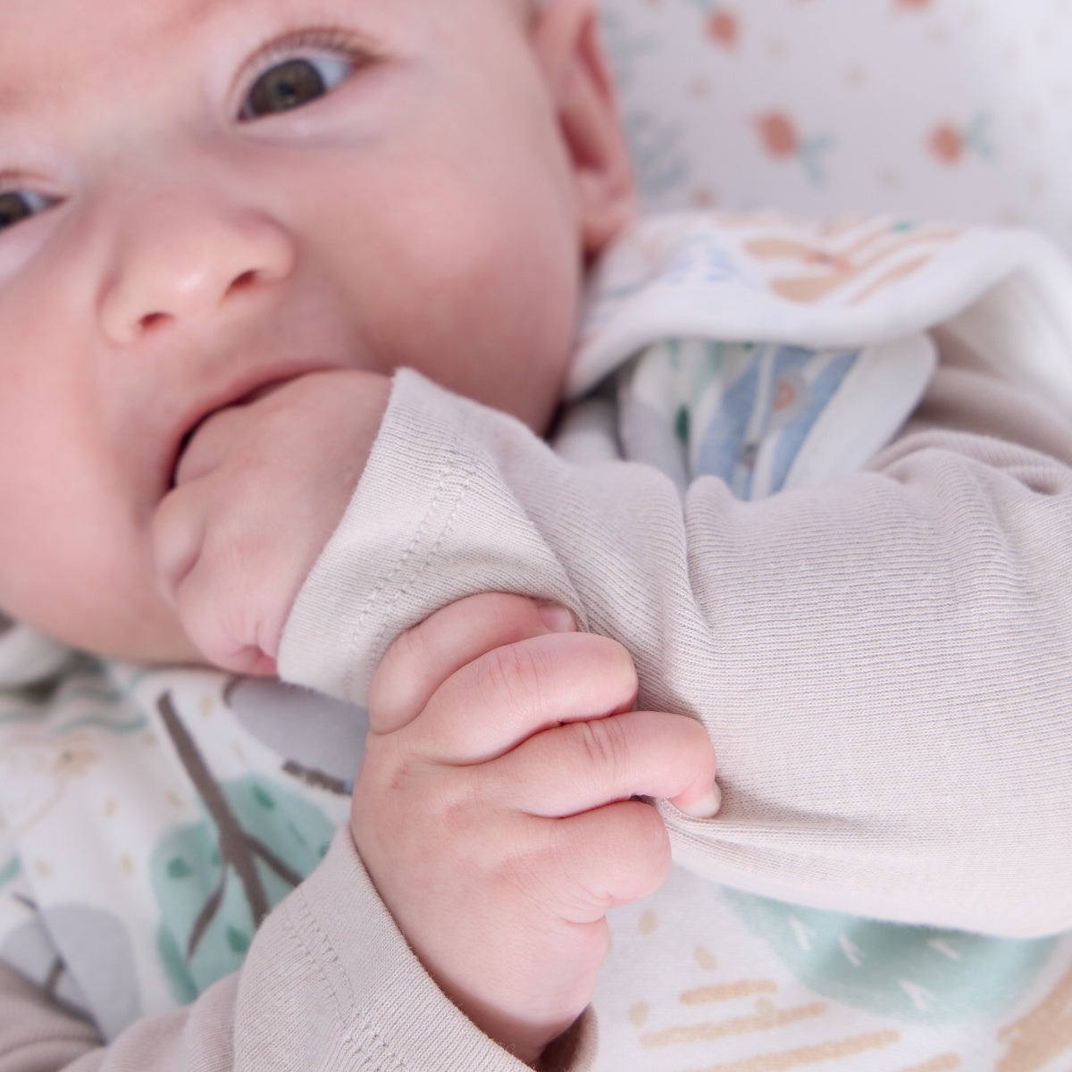 Close-up of baby wearing the Good Life sleep bag, gently chewing sleeve while lying on matching countryside themed sheet