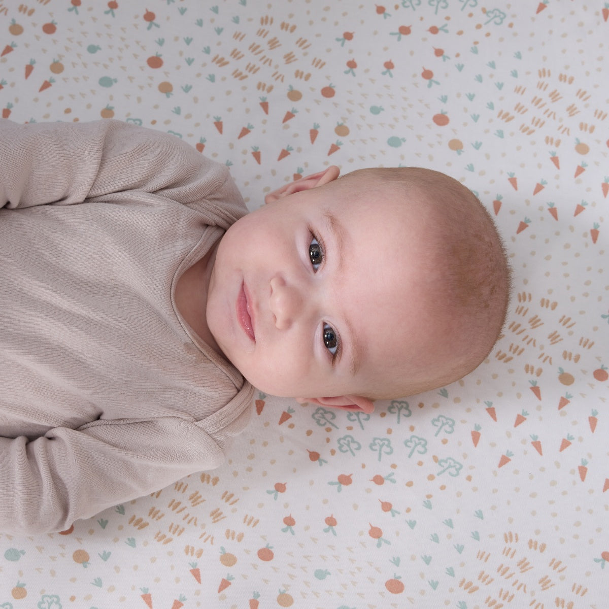 Close-up of baby smiling on the Good Life fitted sheet in cream with vegetable prints, blending warmth and whimsical character