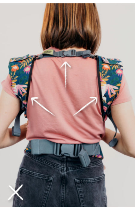 Back view of a woman wearing the Boba X baby carrier, showing strap extension arrows across the shoulder and back area. The image illustrates how to adjust the back strap for optimal comfort and ergonomic fit.