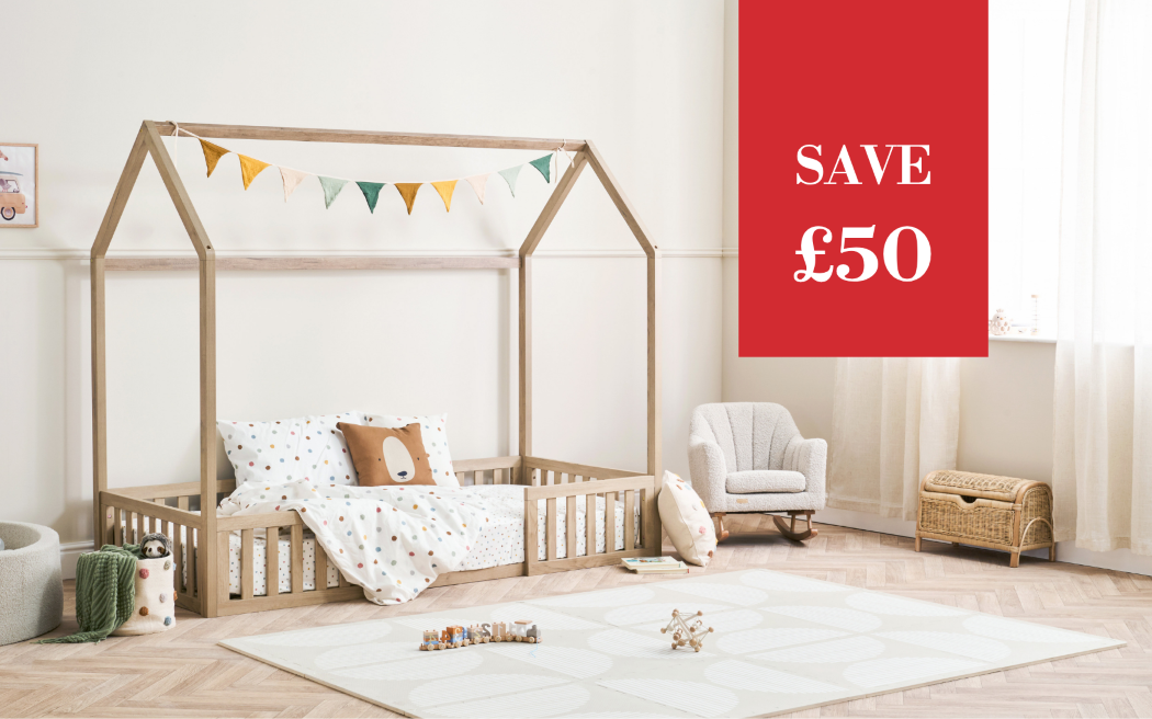 A wooden house-shaped children's bed frame with a canopy, styled in a bright nursery with white patterned bedding, matching pillows, a small armchair, and a playmat. A large red promotional overlay in the top right reads 'SAVE £50'.