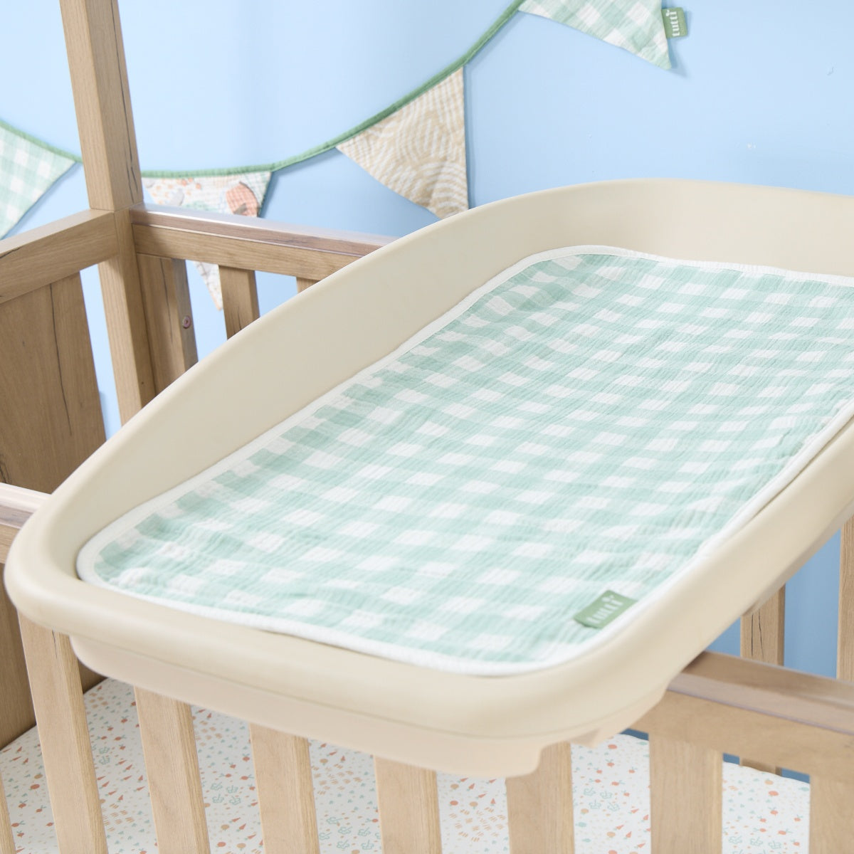 Soft muslin changing mat liner in green gingham print placed on a cot-top changer, adding a calm, coordinated touch to the nursery