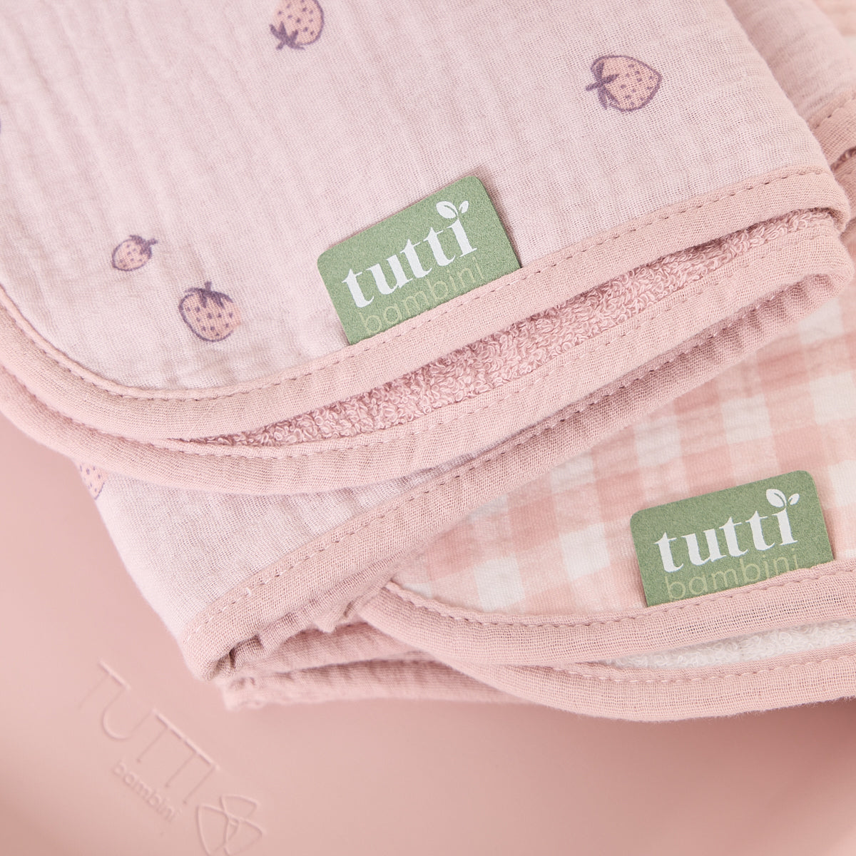 Close-up of soft muslin changing mat liners in pink strawberry and gingham prints, showing Tutti Bambini labels and textured edges