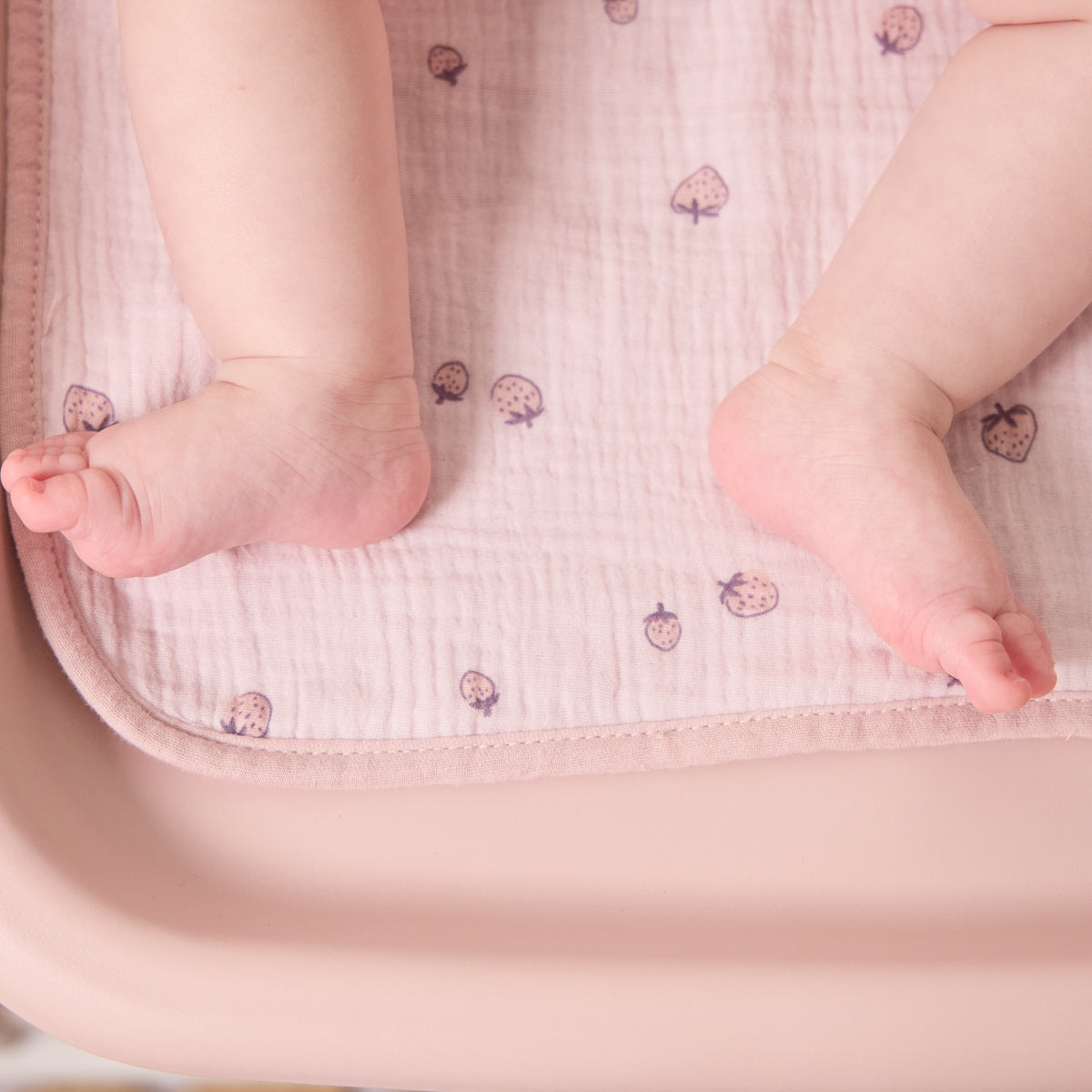 Baby’s feet resting on soft muslin changing mat liner in pink strawberry print, highlighting its gentle, breathable fabric and comfort