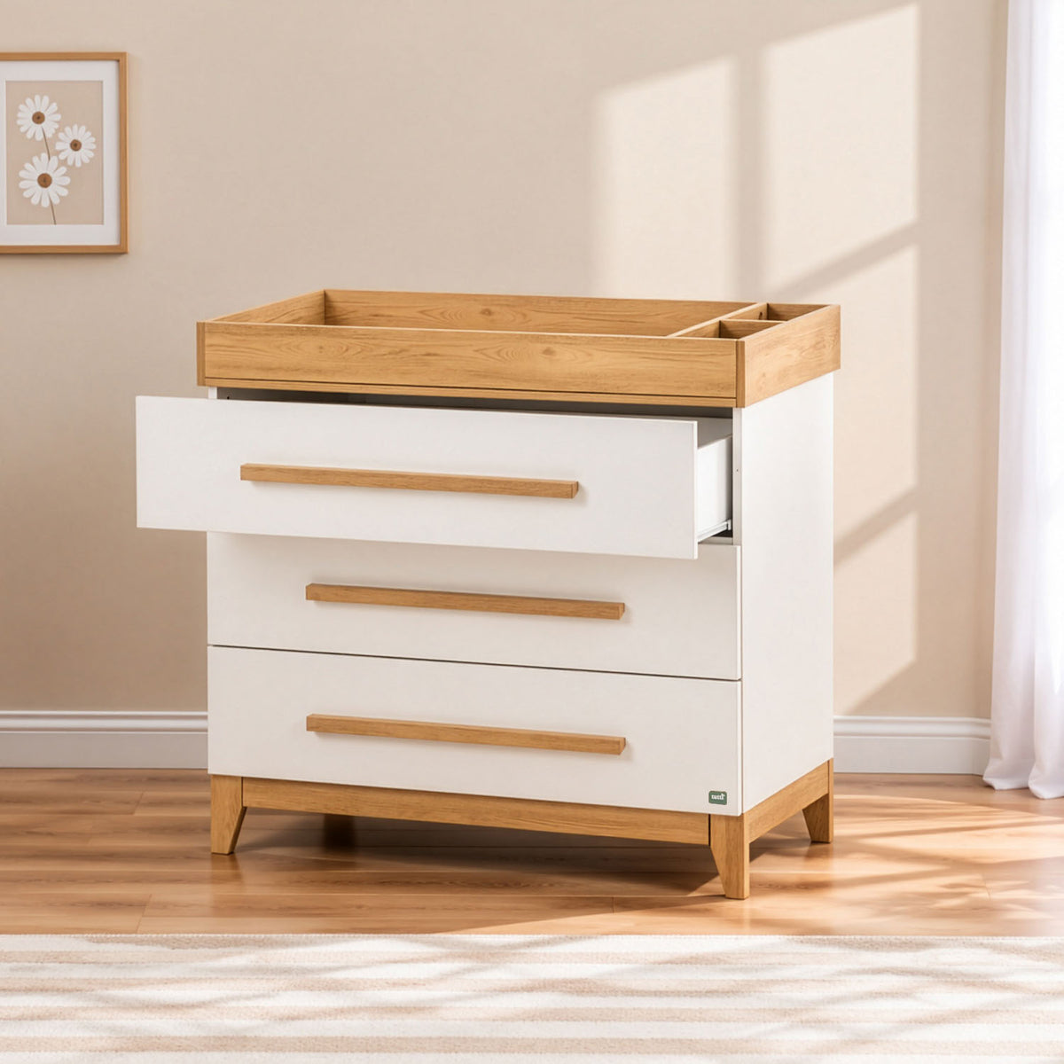 A three-drawer chest of drawers with a golden oak wood top integrated baby-changing unit and matching wood drawer pulls, set against a beige wall with a window light casting soft shadows. The top drawer is open.