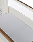 A white bedside crib fitted sheet covering a mattress inside a rich walnut and cashmere vintage stripe crib.