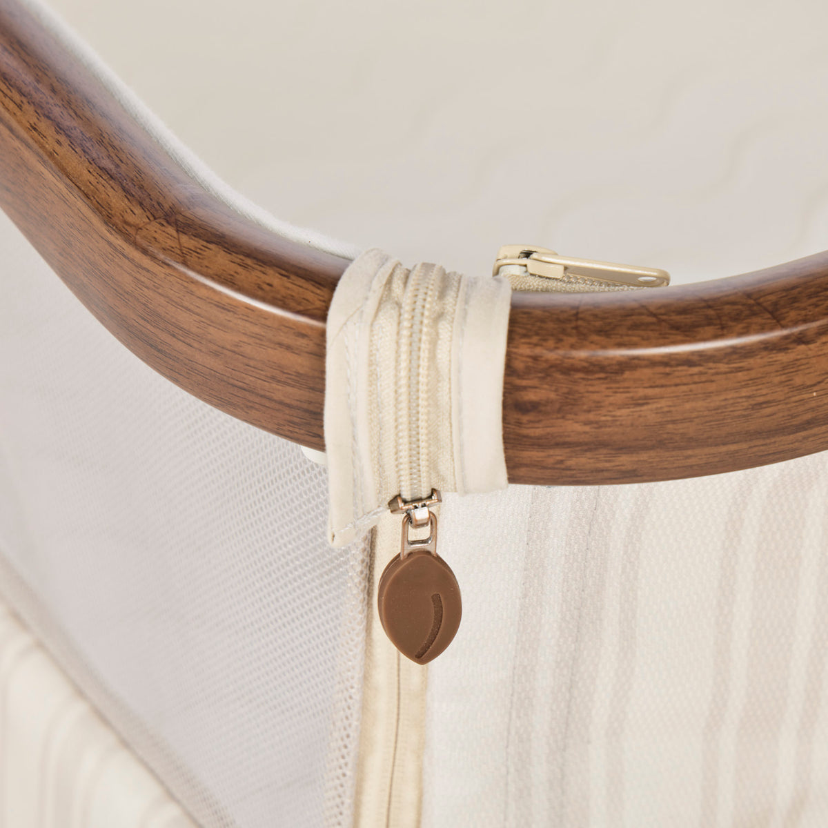 A close-up of a cream-colored mesh bedside crib with a wooden curved frame, featuring a beige zipper pull tab with a brown leaf-shaped charm representing Origin.