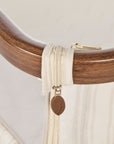 A close-up of a cream-colored mesh bedside crib with a wooden curved frame, featuring a beige zipper pull tab with a brown leaf-shaped charm representing Origin.