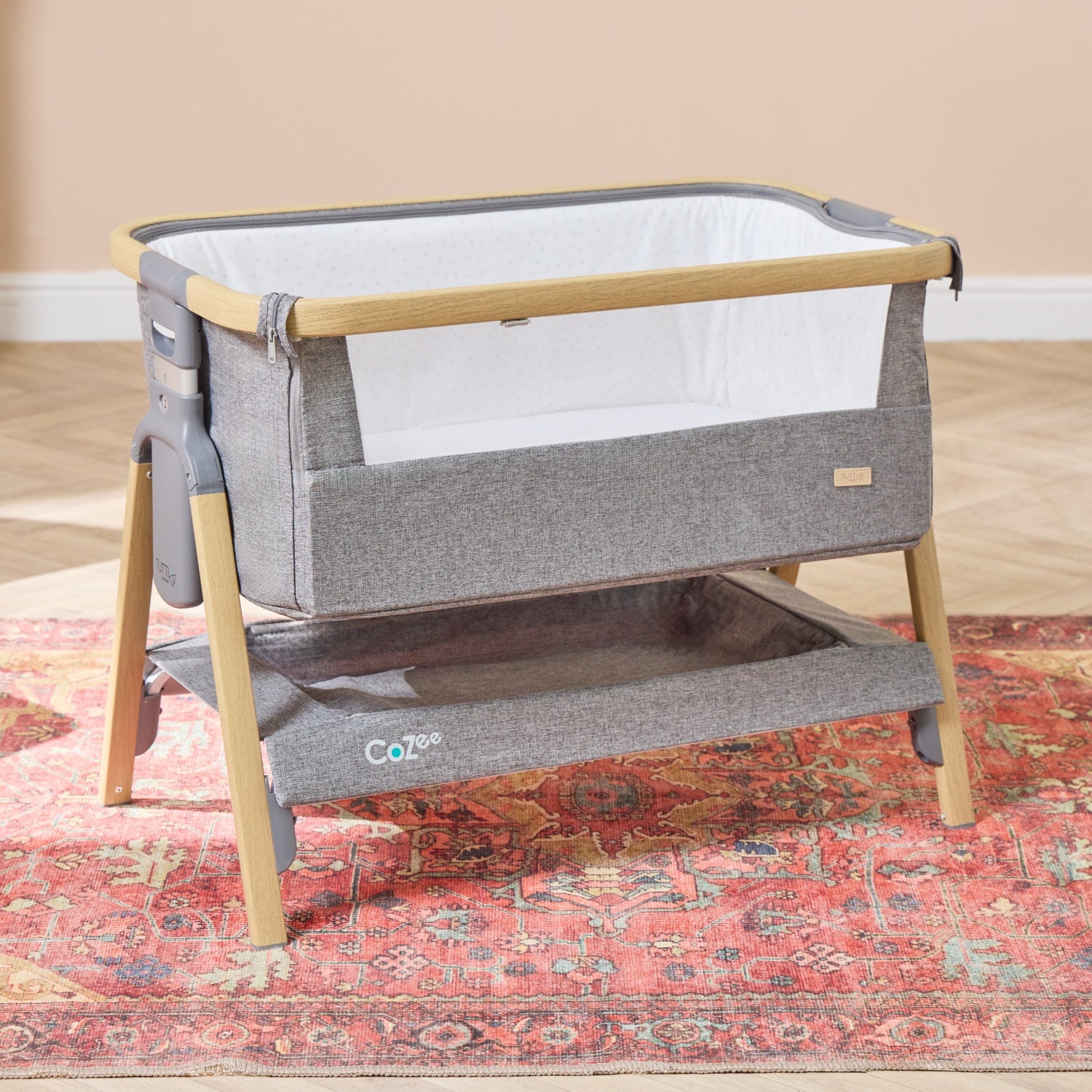 CoZee bedside crib in oak and charcoal shown in a bright nursery, highlighting its breathable mesh sides and handy storage shelf beneath