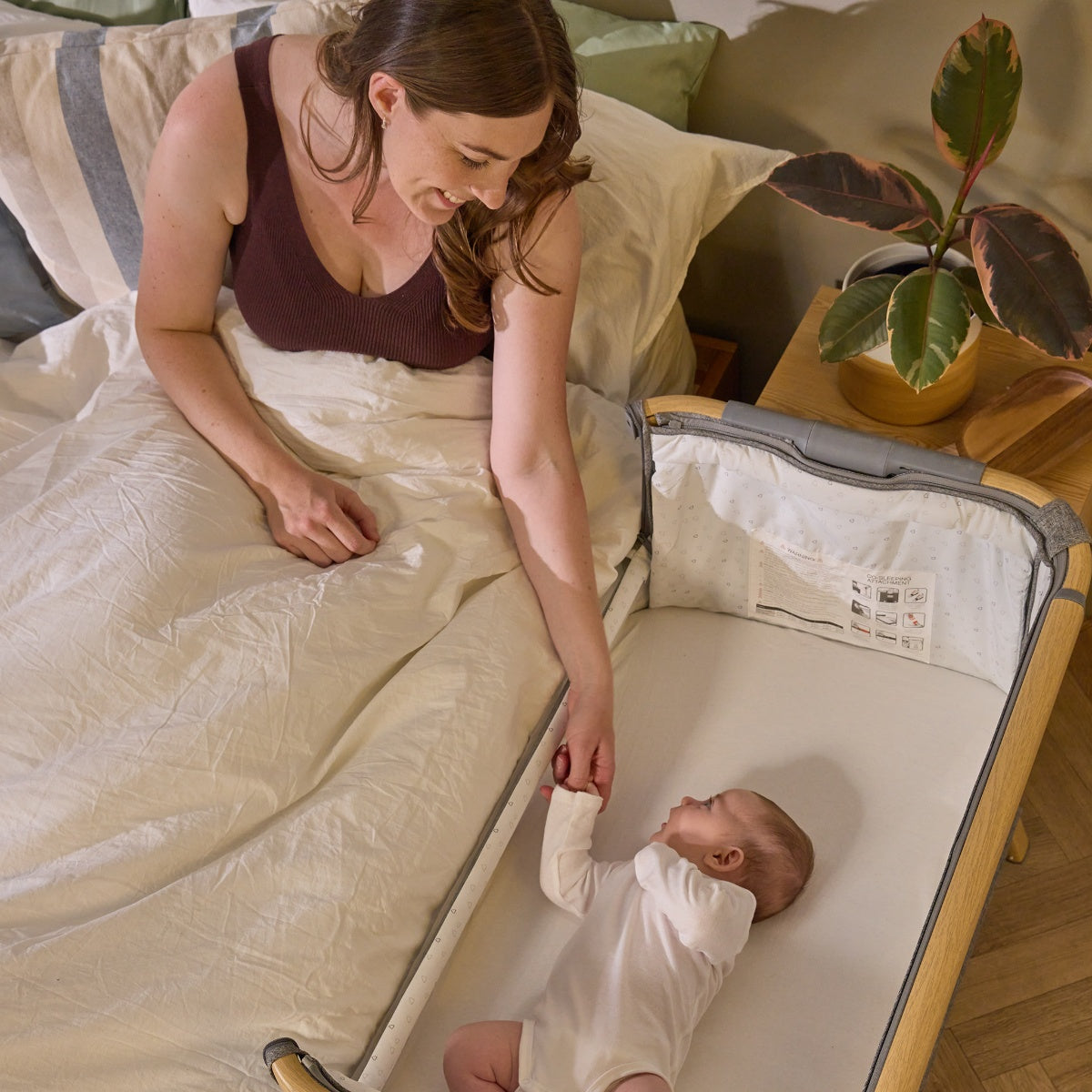 Mother holds baby’s hand while lying beside the CoZee bedside crib in oak and charcoal, highlighting safe co-sleeping design