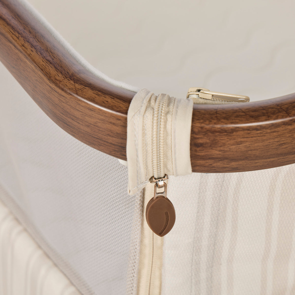 A close-up of a cream-colored zipper with a brown leaf-shaped pull tab, representing Origin, attached to a curved dark brown wooden frame, with a white mesh panel below.