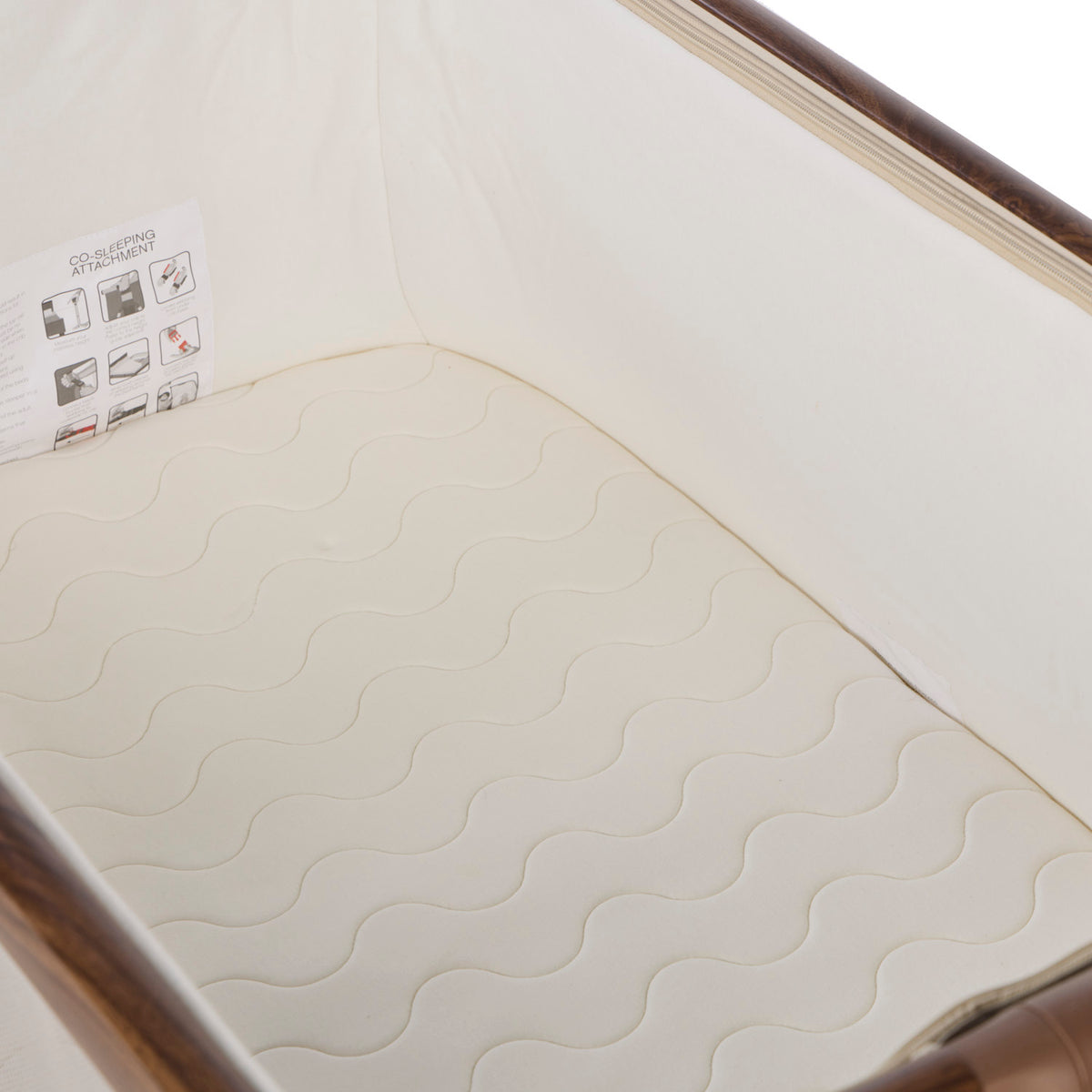 A close-up of a cream-colored mattress with a wavy quilted pattern, fitted inside a bedside crib with a white fabric liner and wooden rails.