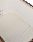 A close-up of a cream-colored mattress with a wavy quilted pattern, fitted inside a bedside crib with a white fabric liner and wooden rails.
