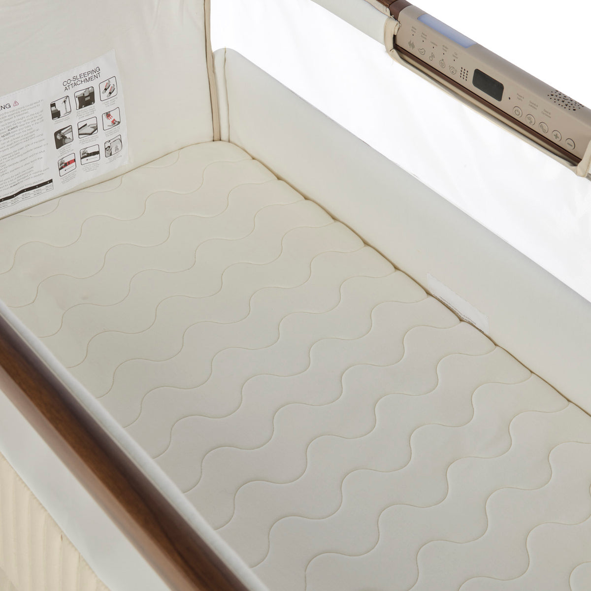 A close-up of a cream-colored mattress with a wavy quilted pattern, shown inside a bedside crib. A control panel is visible on the side rail, and an instruction sheet is attached to the inner wall.