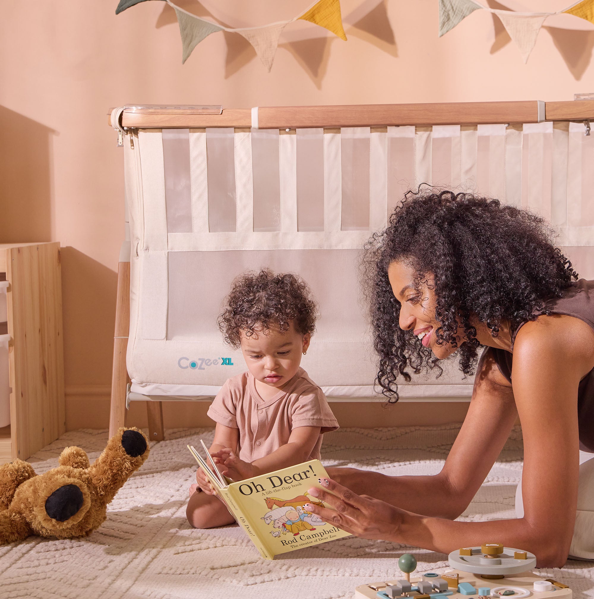 Mum and toddler read together beside the CoZee XL Scandi Cot, showing its mesh sides and versatile design