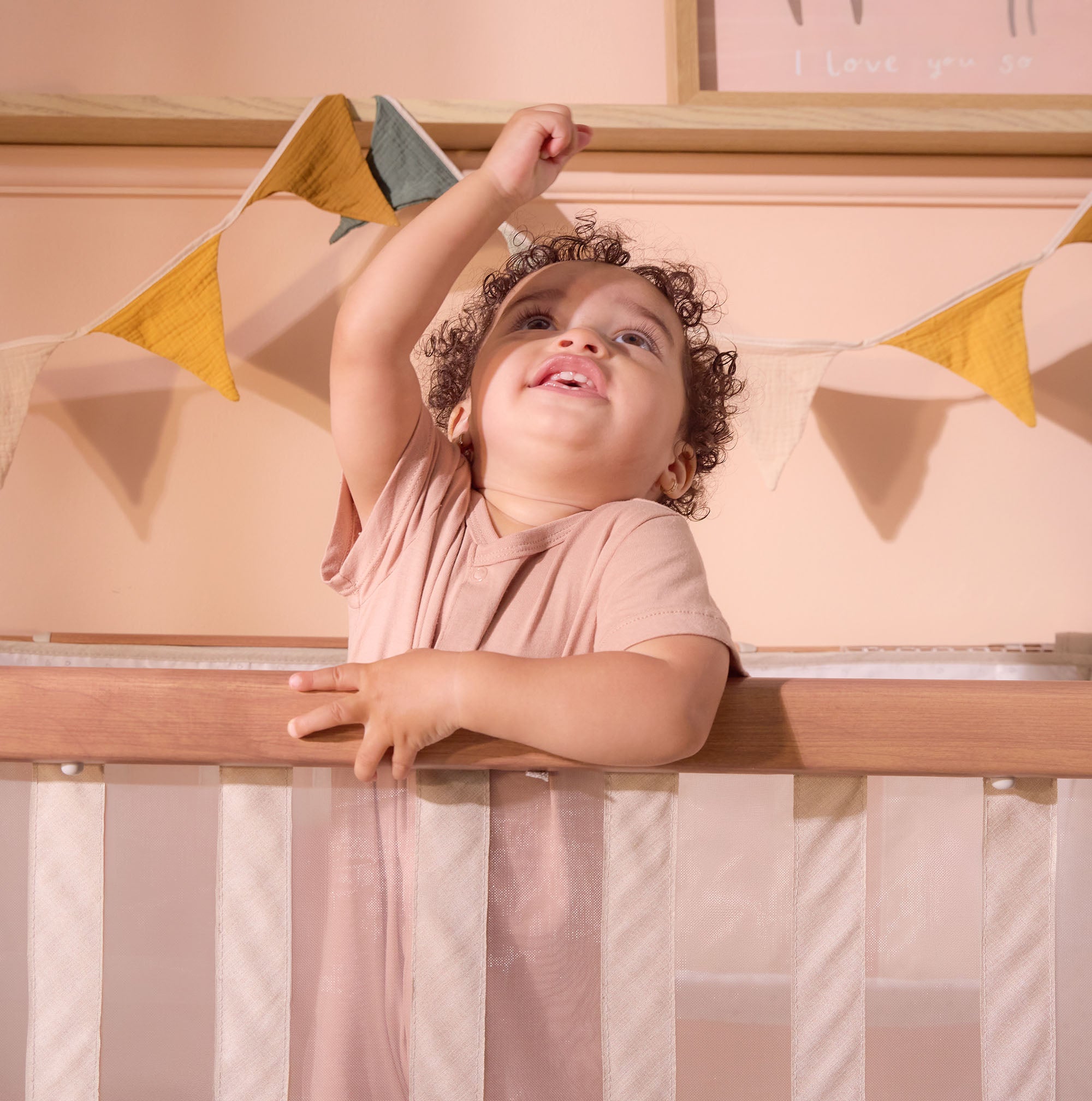 Smiling toddler stands in the CoZee XL Scandi Cot, holding the rail and reaching up under nursery bunting
