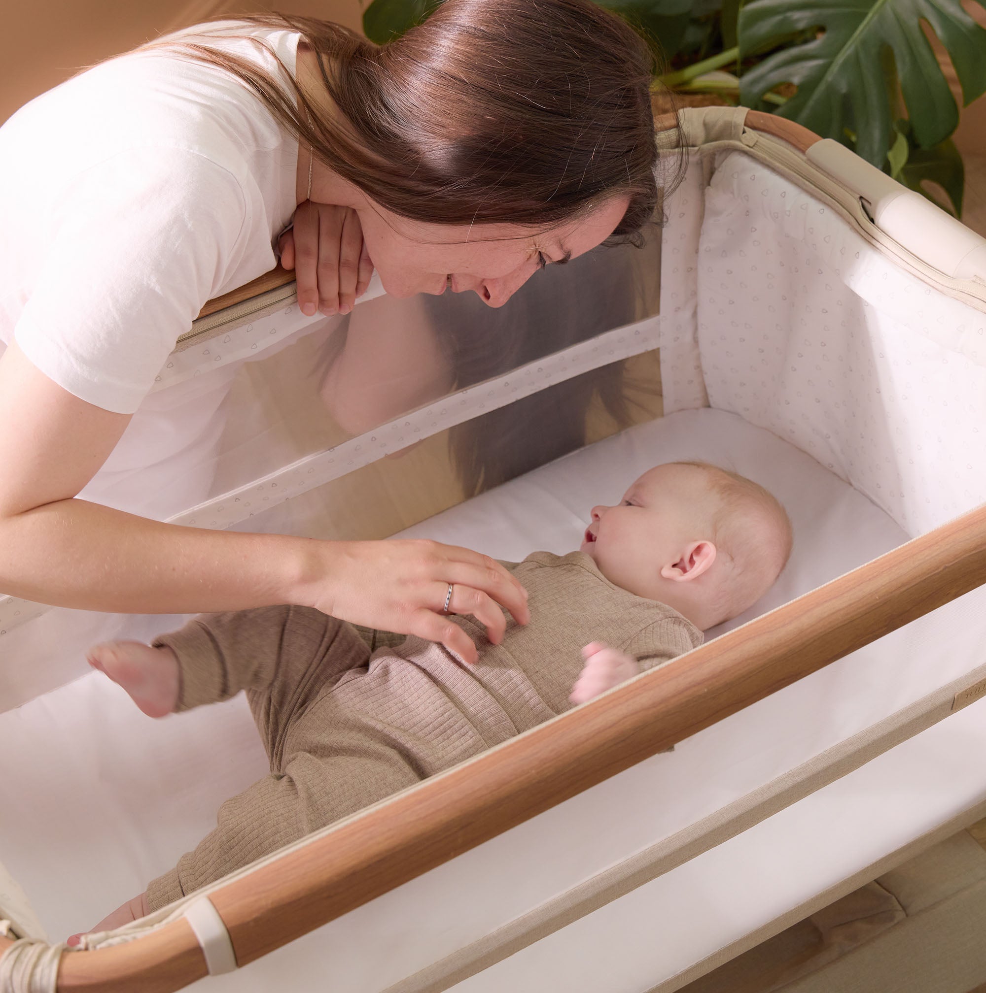 Mum smiles while tickling her baby lying in the CoZee Scandi bedside crib, highlighting mesh sides, breathable fabric, and accessible co-sleeping design