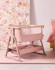 Angled lifestyle view of the CoZee Air 2 bedside crib in blush, highlighting rocking legs and soft fabric finish.