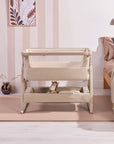 Lifestyle view of the CoZee Air 2 in cocoa beside the bed, showing breathable mesh sides and integrated storage basket below.