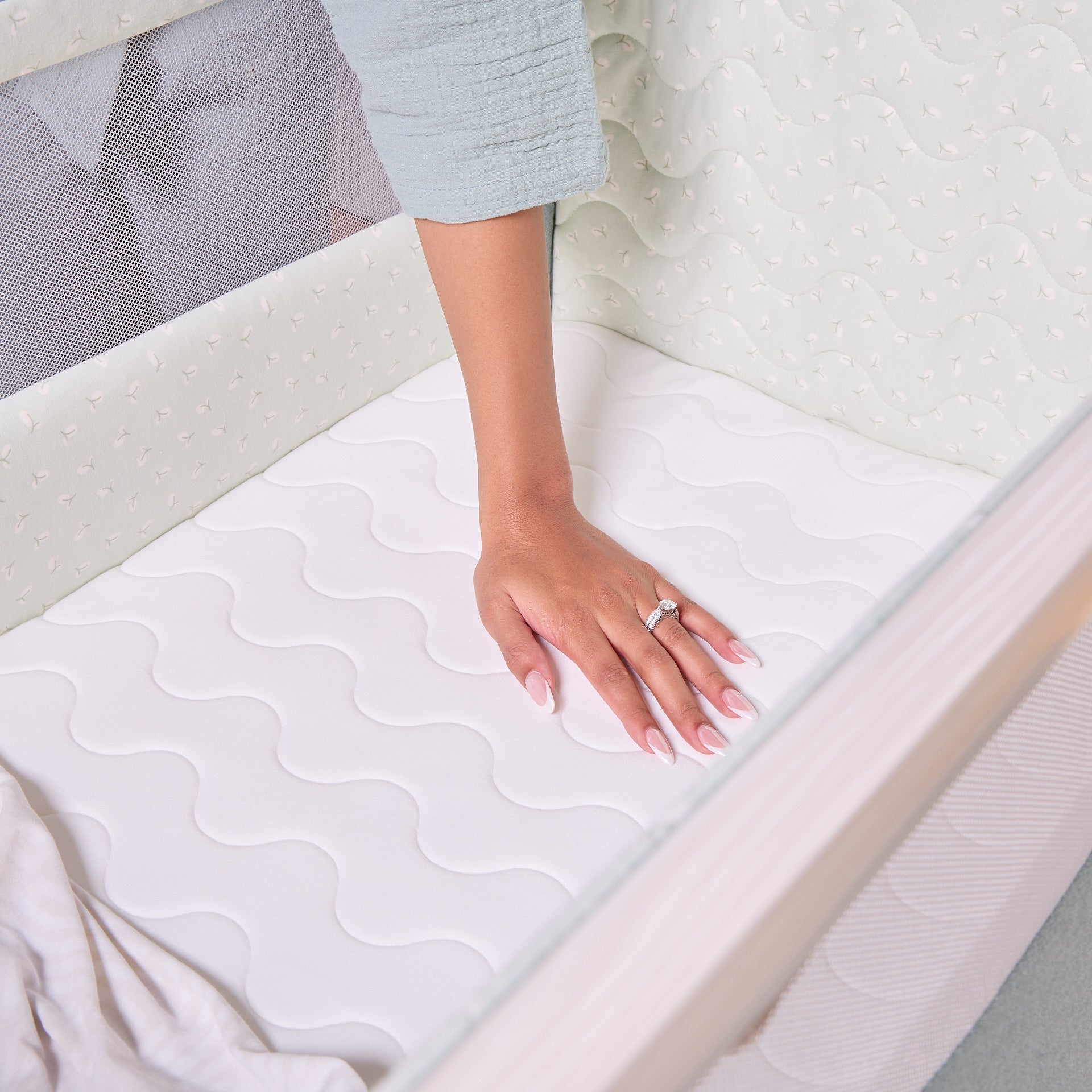 Hand pressing the breathable mattress inside the CoZee Air 2 bedside crib in kelp and whitewash, highlighting comfort and support.