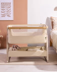 Front view of the CoZee Air 2 bedside crib in toasted oak/sand styled beside the bed, showing breathable mesh sides and storage shelf.