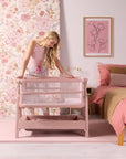 Mother leaning over CoZee Zen bedside crib in blush beside bed, styled in soft pink bedroom with floral wallpaper.