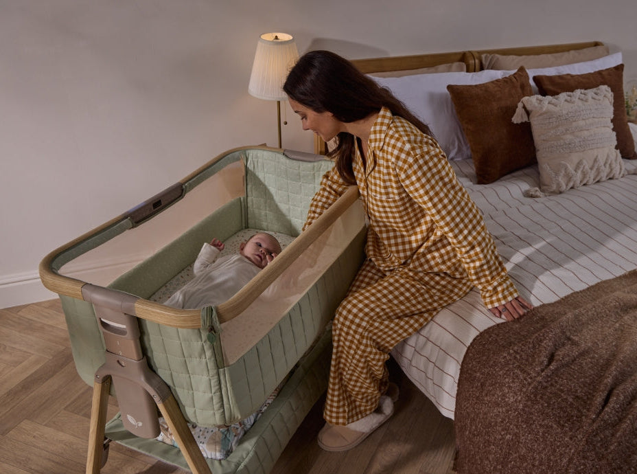 Mother seated beside CoZee sense bedside crib, gently soothing baby through breathable mesh panel.