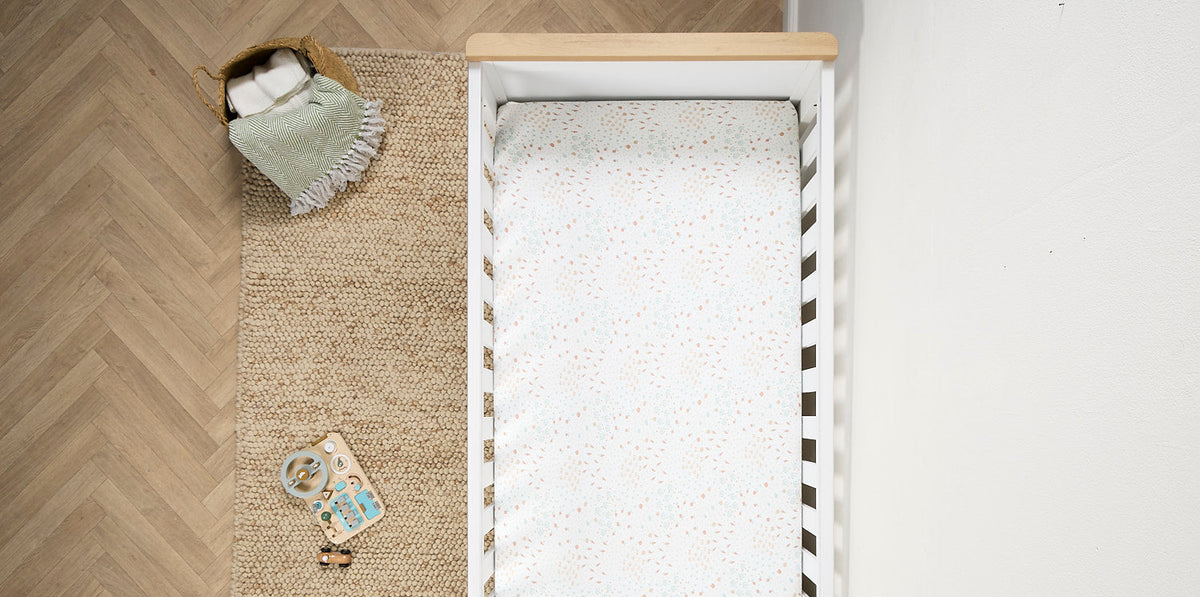 Top-down view of a white baby cot bed with a speckled white mattress and fitted sheet, placed on a woven rug beside a light wood floor and a small blanket.