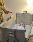 Parent activating soothing sensory light bar on CoZee Sense bedside crib in soft walnut and cocoa while baby lies inside.