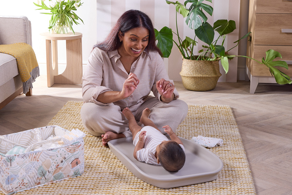 Mum sits with baby on Cushi Mini changing mat in mushroom on rug, showing portable design, wipe-clean finish, and cosy setting.