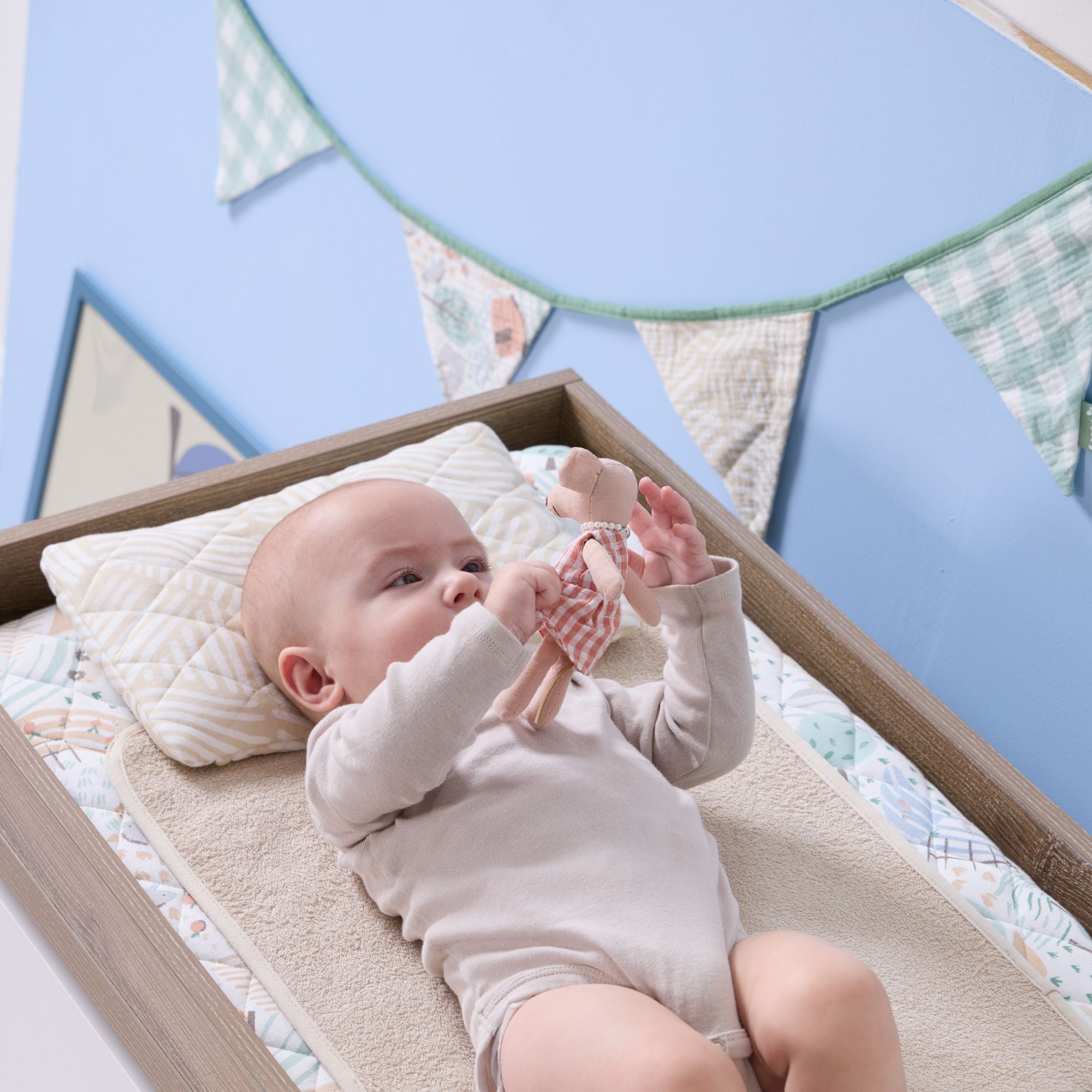 Baby lying on a changing mat with woodland print cover, reaching for a soft toy beneath green gingham nursery bunting.