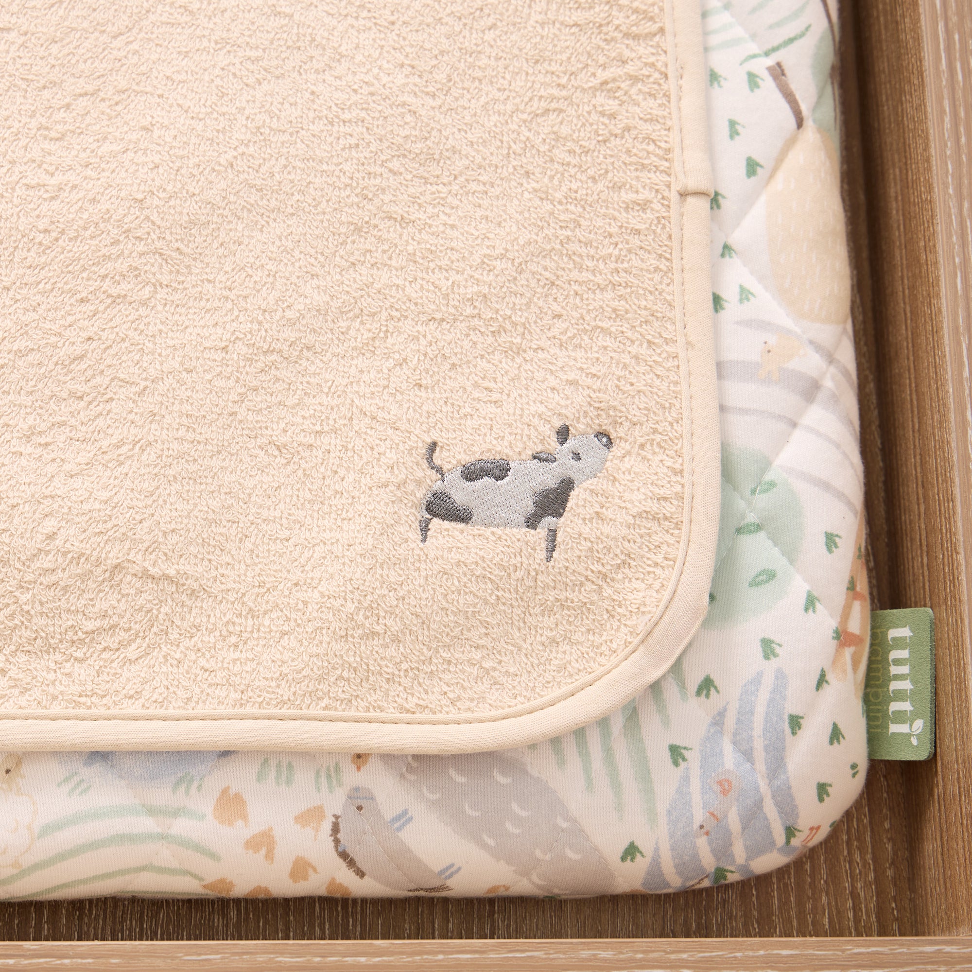 Close-up of the Good Life luxury changing mat with a cow illustration and Tutti Bambini tag on a wooden surface.