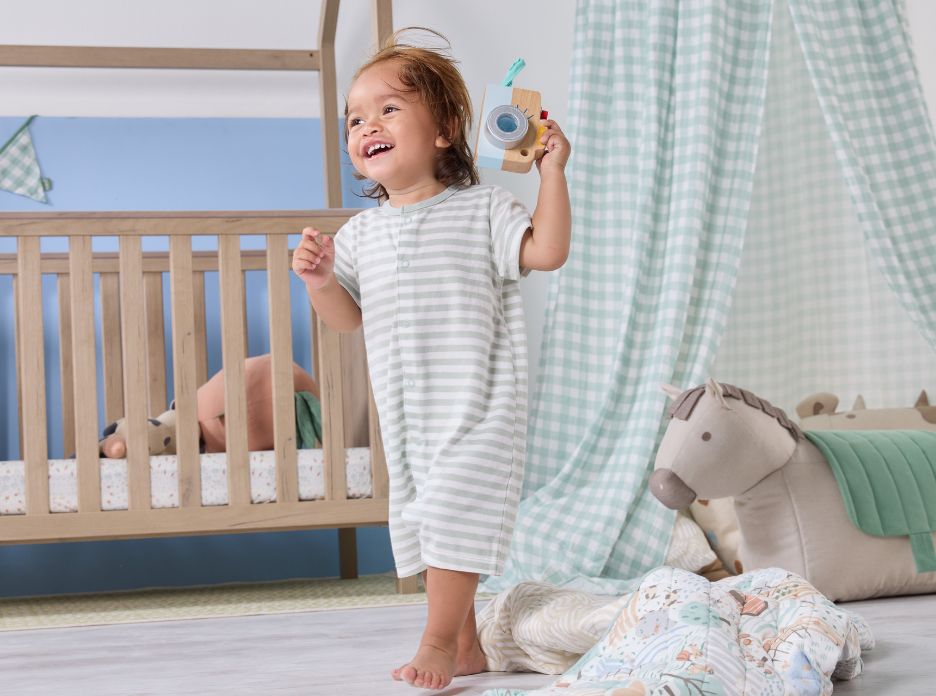 Toddler standing and smiling in a styled nursery with wooden cot and green good life nursery canopy, creating a playful, cosy den space.