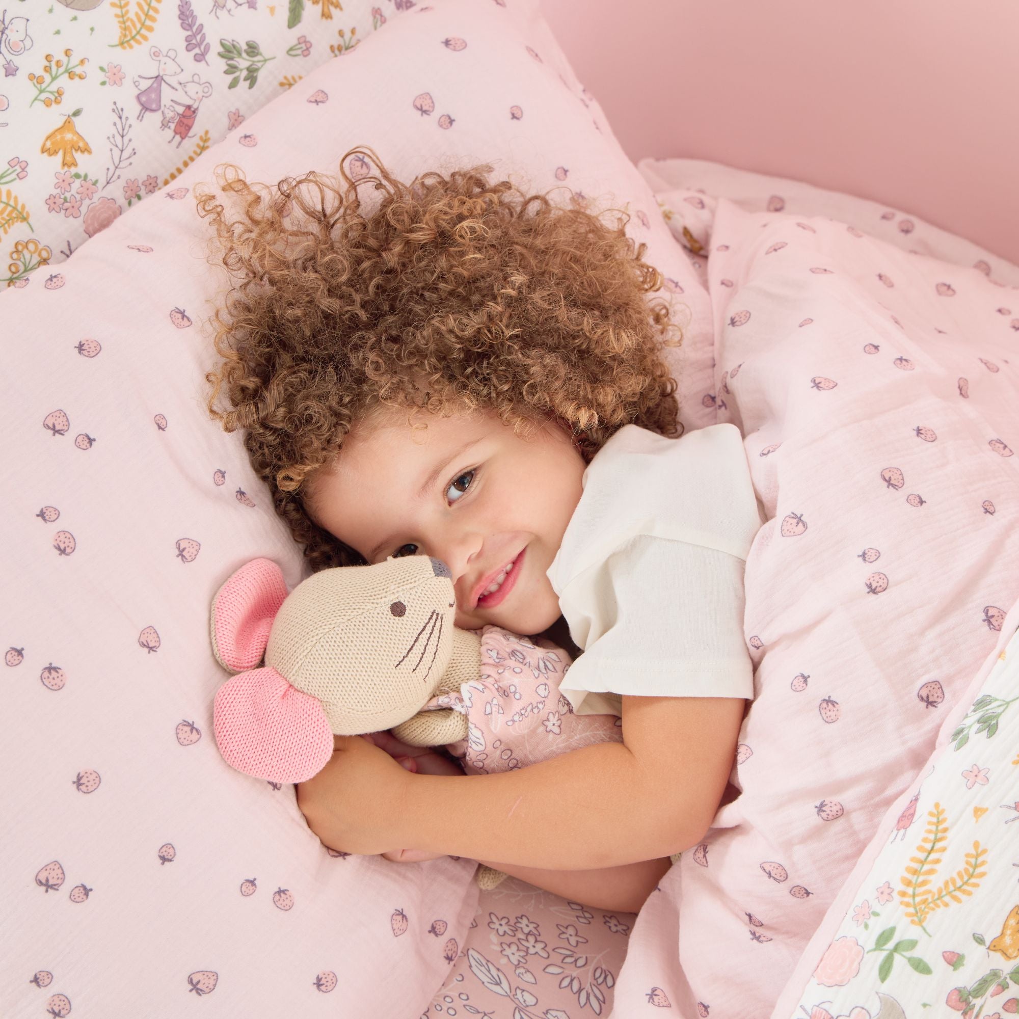 Child lying in bed with Margot Mouse knitted toy, surrounded by pink bedding and floral patterns.
