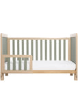 A Scandi-style cot bed with rounded corners, sturdy block legs, sage weave texture on the ends and slats with a matching bed guard.