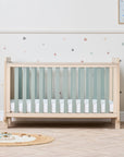 A Scandi-style cot bed with rounded corners, sturdy block legs, sage weave texture on the ends and slats, and a pickled oak frame.