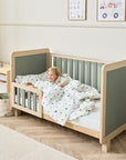 A child laying on a Scandi-style cot bed with rounded corners, sturdy block legs, sage weave texture on the ends and slats, and a matching bed guard.