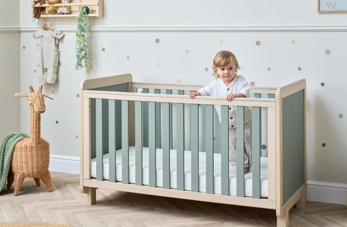 Toddler standing inside the Kuba cot bed in pickled oak and sage weave, highlighting sturdy slats, calm tones and supportive cot design