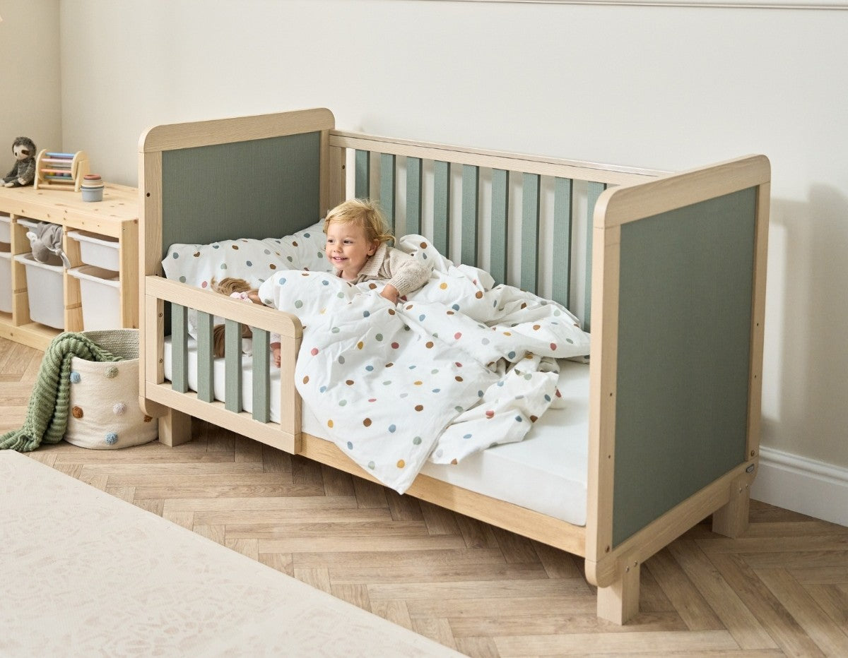 Toddler resting comfortably in the Kuba cot bed in pickled oak and sage weave, showing low toddler rail and spacious sleep surface