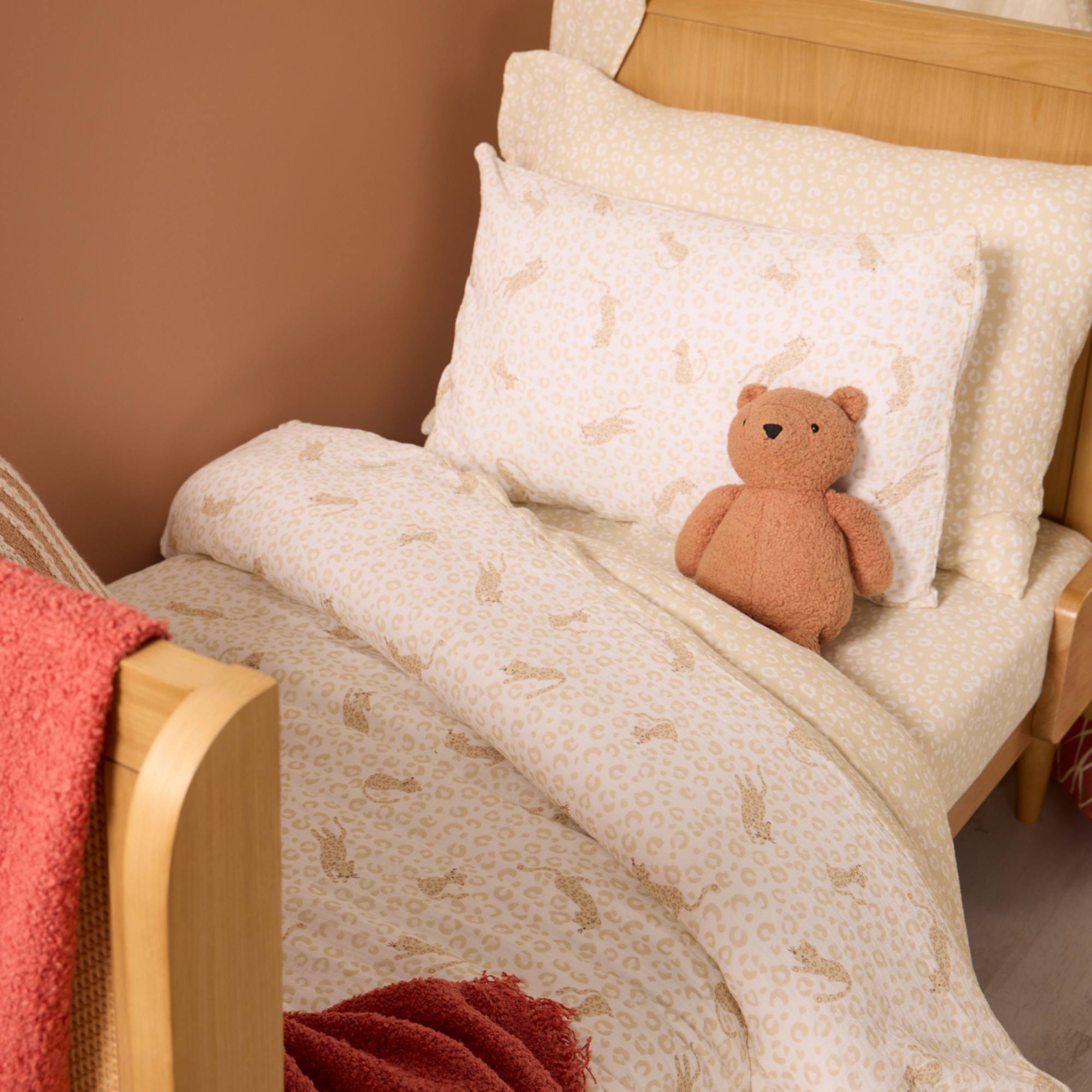 Children's bed with Love Cats patterned bedding and a teddy bear, against a brown wall.