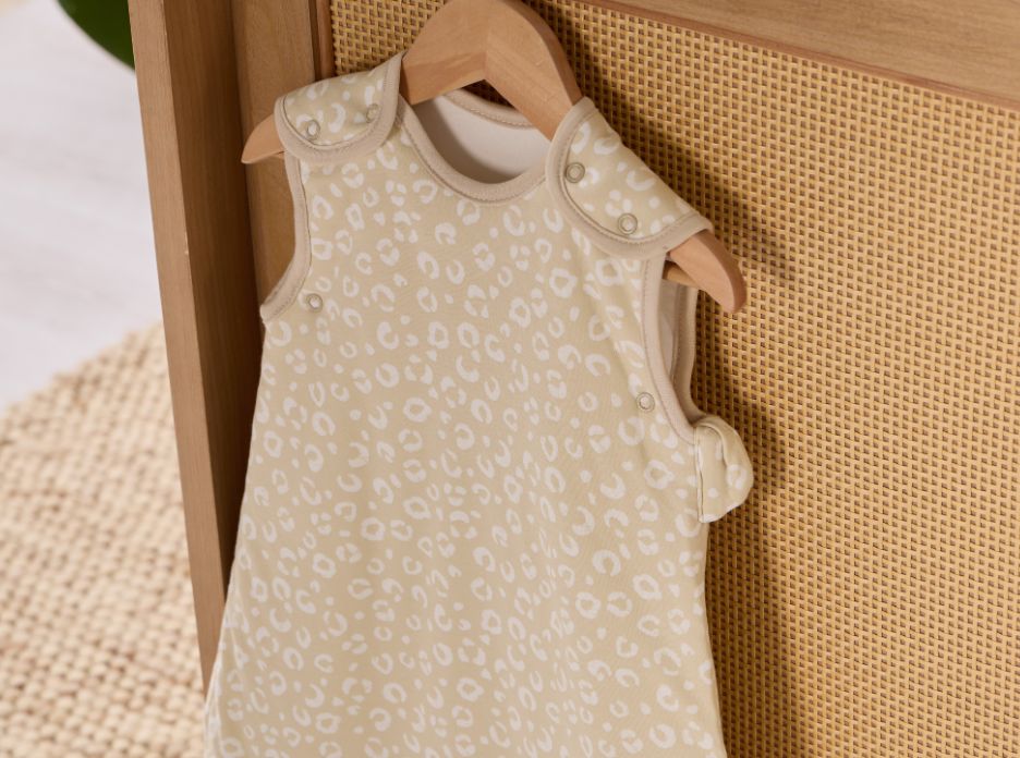 Baby sleep bag with white animal print pattern on a wooden hanger against a rattan and wood cot bed.
