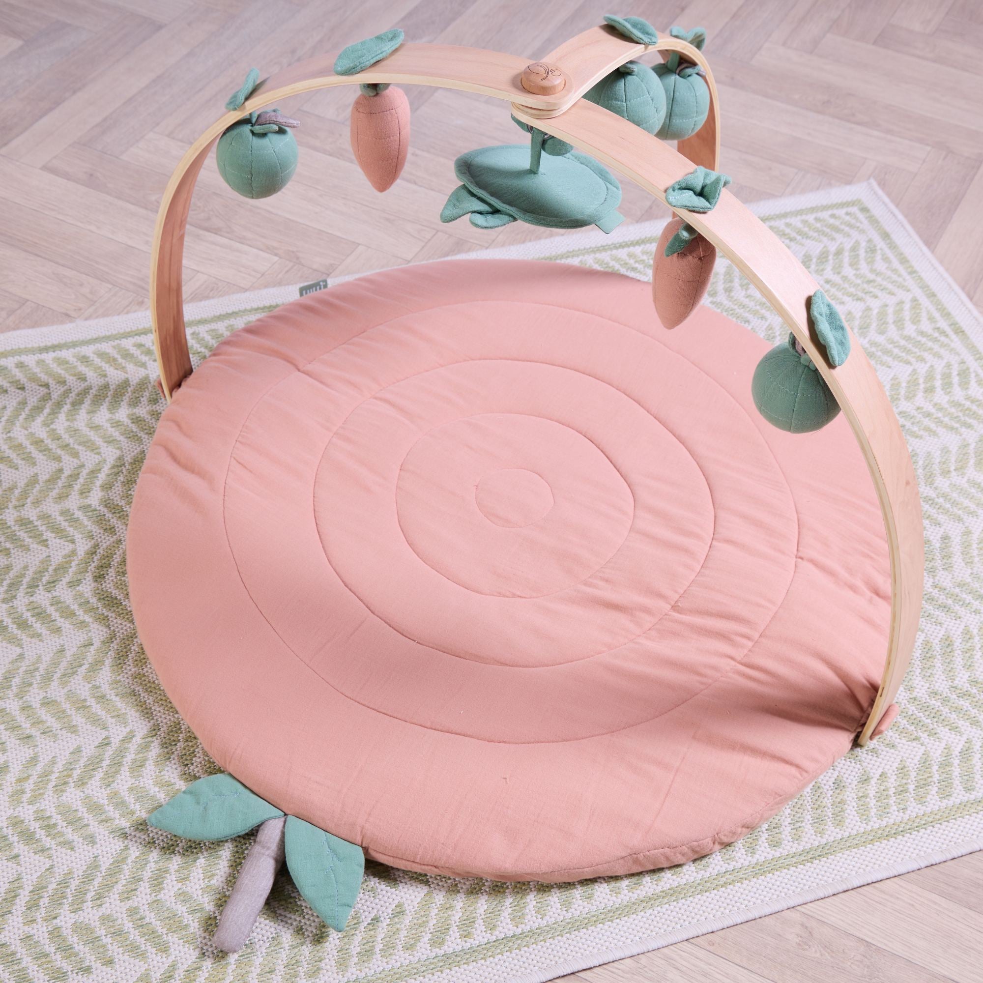 Angled view of good life muslin baby playmat with wooden arch and hanging fruit toys on woven rug.