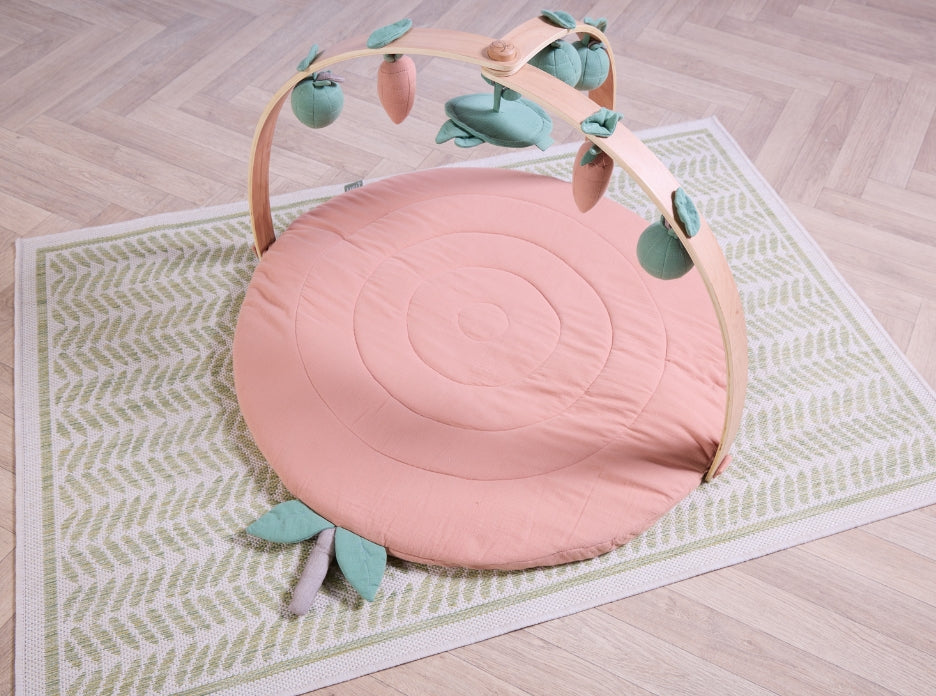 Round terracotta muslin padded playmat with wooden arch and hanging fruit toys on patterned rug.